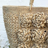 Tasken er lavet i lys natur raffia og har store formede blomster på fronten. HAr flettede raffia hanke