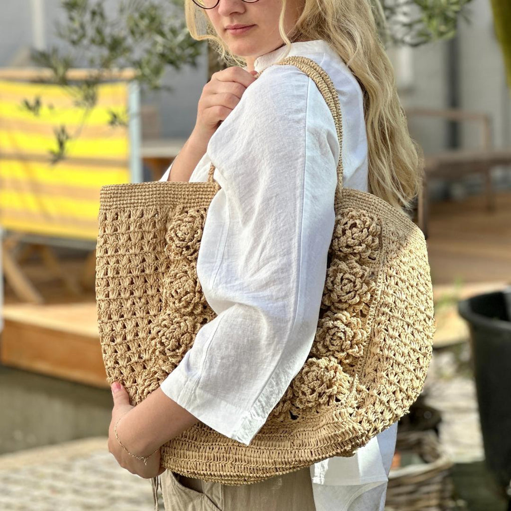 Tasken er lavet i lys natur raffia og har store formede blomster på fronten. HAr flettede raffia hanke
