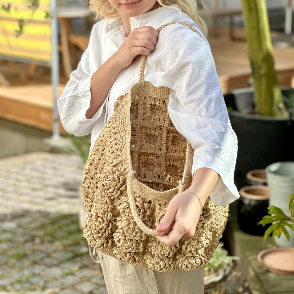 Tasken er lavet i lys natur raffia og har store formede blomster på fronten. HAr flettede raffia hanke