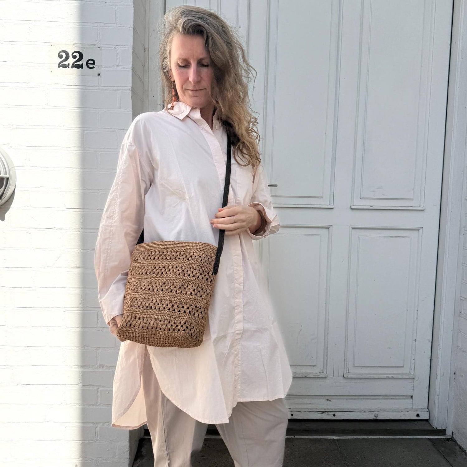 Flettet skuldertaske i brun naturfarve med lang rem båret crossbody til lys skjorte og bukser