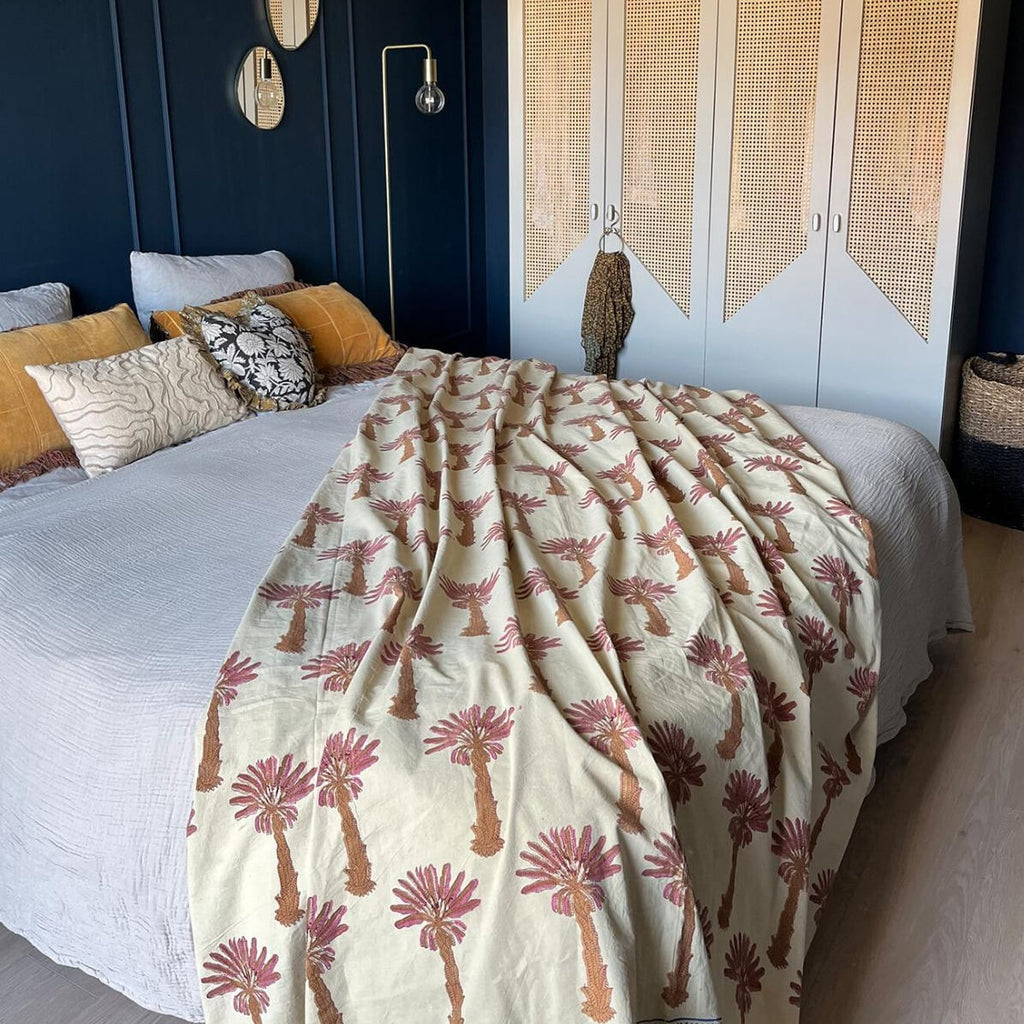 Et dug/sengetæppe med et mønster af brune og lyserød palmer på en beige baggrund i midten og en blå og gul mønstret kant