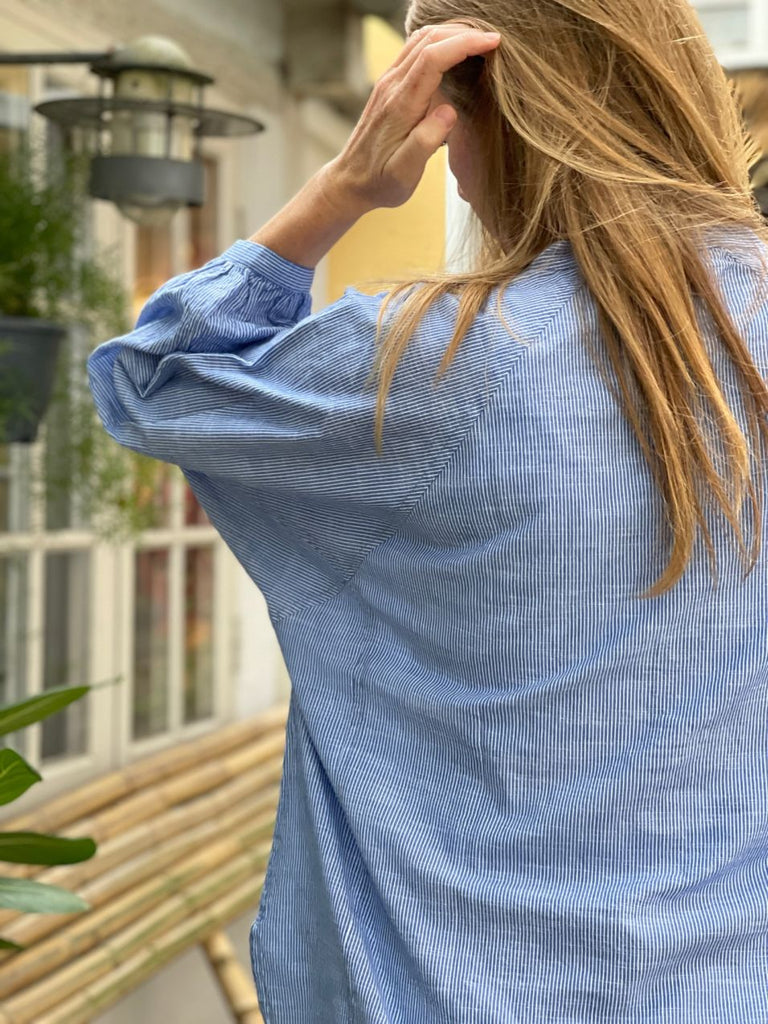 Stribet lyseblå oversized skjorte med ballonærmer og kinakrave. Den løse 'Tokyo' skjorte har en løs og rummelig pasform. Skjorten er gennemknappet og er lavet i en let økologisk bomuldspoplin, som gør skjorten åndbar og behagelig at have på. Skjorten er one size og passer str 34-44.